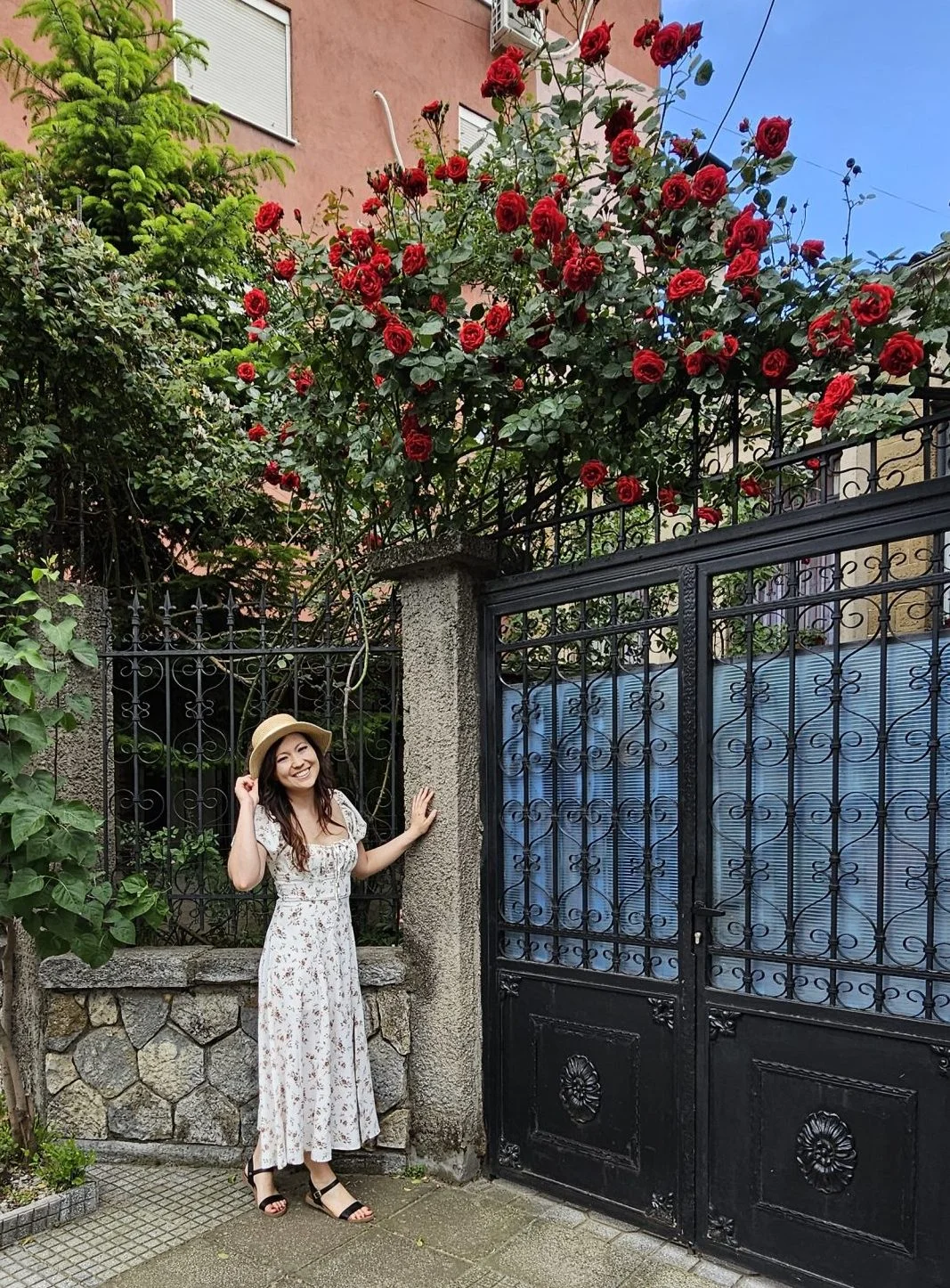 About Woman in a white dress and wearing a hat standing in front of a door fence with roses in Korca Albania